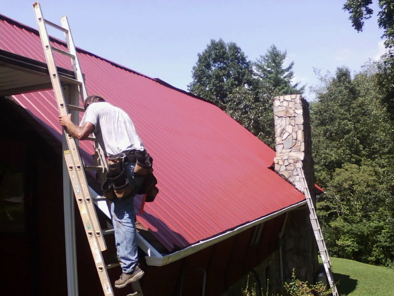 Roofer on a ladder beside a red metal roof and stone chimney — Soffit & Fascia Repair by Roofing Brothers in Port Republic